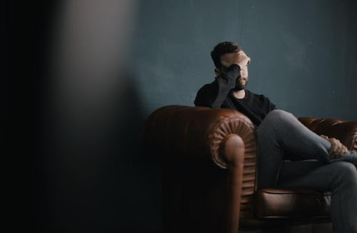 Photo of a man sitting on a brown leather couch and holding his hand on his head